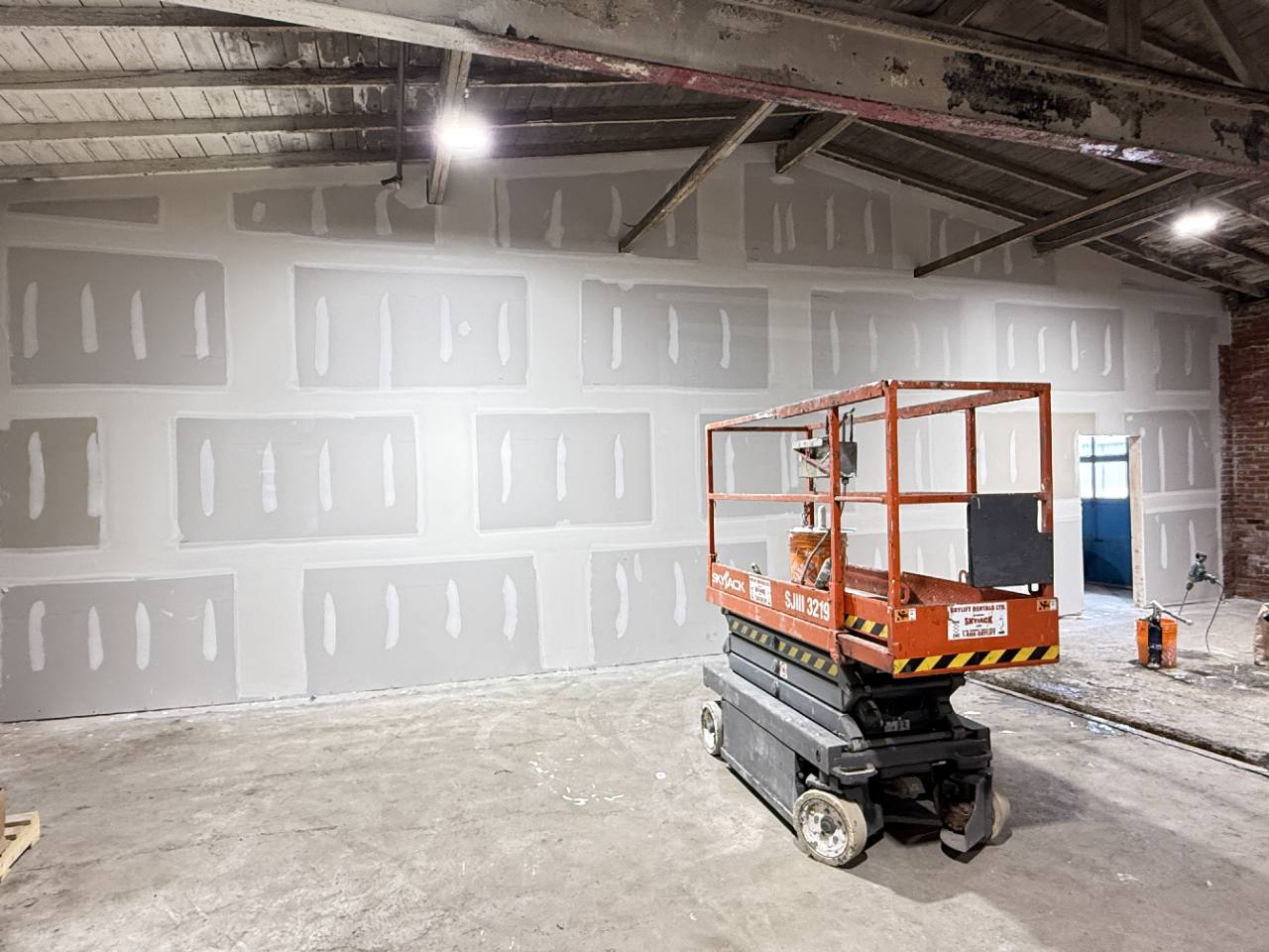 Taped drywall joints and fasteners across a tall wall, with a scissor lift used during the work.